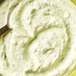 Small bowl of creamy garlic lime crema with visible green cilantro flecks, garnished with fresh cilantro leaves and lime wedges on a rustic wooden table.