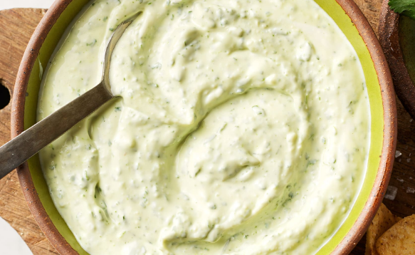 Small bowl of creamy garlic lime crema with visible green cilantro flecks, garnished with fresh cilantro leaves and lime wedges on a rustic wooden table.