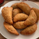 Golden-brown half moon pies on a wooden board, crispy breaded exterior, served with a bowl of garlic lime crema and garnished with fresh cilantro.